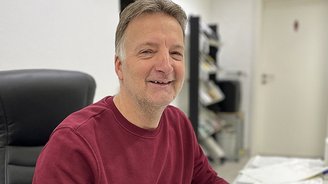 Freundlicher Büroangestellter bei der Arbeit Ein lächelnder Mann sitzt an einem Schreibtisch, umgeben von Unterlagen und einem Stuhl in einem hellen Büro.
