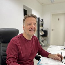 Freundlicher Büroangestellter bei der Arbeit Ein lächelnder Mann sitzt an einem Schreibtisch, umgeben von Unterlagen und einem Stuhl in einem hellen Büro.