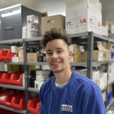 Junger Mann lächelt in einem Lagerraum voller Kisten Ein junger Mann mit lockigem Haar lächelt freundlich vor einem Regal voller bunter Kisten und Kartons.