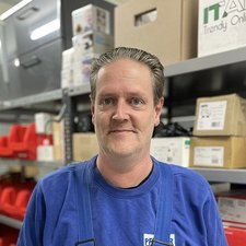 Mitarbeiter in blauer Kleidung vor Lagerregalen In einem Lagerraum steht ein Mann in blauer Kleidung, umgeben von Kartons und roten Behältern, mit einem entspannten Gesichtsausdruck.