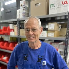 Freundlicher Mitarbeiter vor Regalen mit Lagerwaren Der Mann trägt ein blaues Oberteil und eine Latzhose, während er in einem gut organisierten Lager steht.