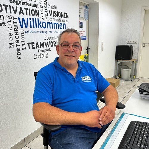 Ein lächelnder Mann in einem blauen Poloshirt sitzt an einem Schreibtisch in einem modernen Büro mit inspirierenden Wänden.