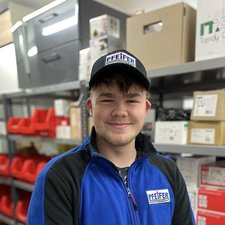 Lagerarbeiter mit Pfeiffer-Mütze in einem Lagerraum Der Mitarbeiter trägt ein blaues Oberteil mit dem Logo 'PFEIFFER' und steht in einem Lager mit roten Aufbewahrungskisten im Hintergrund.