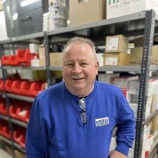 Freundlicher Mitarbeiter in blauer Kleidung im Lager Ein lächelnder Mann in blauer Kleidung steht in einem Lager mit roten Regalen und Kartons im Hintergrund.