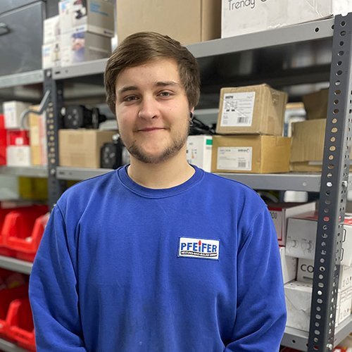 In einem gut organisierten Lager lächelt ein junger Mann in einem blauen Sweatshirt mit dem Logo 'PFEIFFER'.