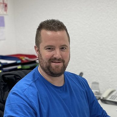 Ein Mann mit Bart sitzt an einem Schreibtisch, lächelt freundlich und trägt ein blaues T-Shirt in einem hellen Büro.